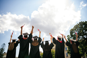 Rear portrait of group students raise fists hold diploma mortarboard gown graduate college building...