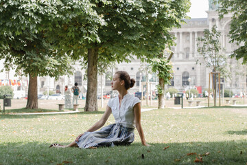 The girl sits thoughtfully on the grass in the park, dressed femininely and elegantly, looks sophisticated and looks away.