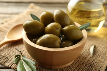 Delicious marinated olives in bowl, green leaves, spoon and oil on table, closeup