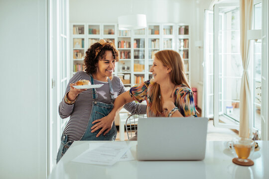 Happy expecting lesbian couple working from home and having snack in cozy home office