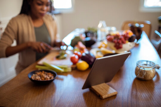 Black woman cooking healthy meal while following recipe on tablet in kitchen with fresh vegetables and fruits - Powered by Adobe