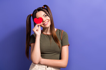 Young woman with playful hairstyle holding a red heart shape while smiling against a purple background