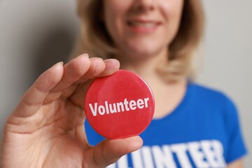 Woman holding button badge with word Volunteer on grey background, closeup