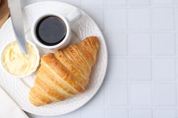 Tasty fresh croissant served with butter and cup of coffee on white tiled table, top view. Space for text
