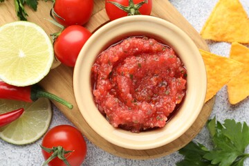 Spicy salsa sauce in bowl, nachos and ingredients on light grey table, flat lay