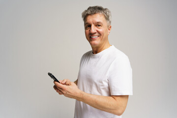 Smiling man in white shirt holds smartphone while standing against plain background indoors