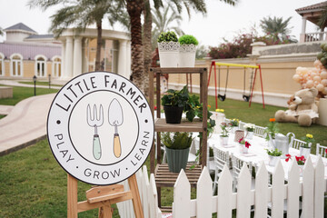 A charming outdoor kids' gardening event, "Little Farmer," with potted plants, a rustic setup, white picket fence, a play area, and decorated tables, fostering hands-on learning in a lush garden setti