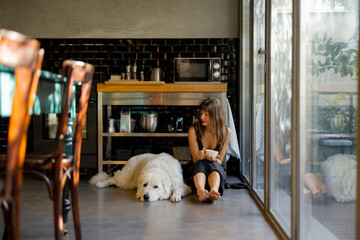 A barefoot woman sits on the floor of a modern kitchen, sipping coffee next to her large white dog....