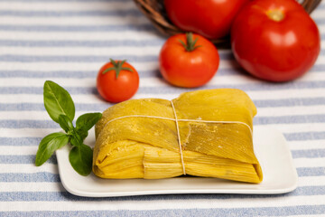 humita envueltas en hojas de choclo cerrada con pitilla sobre un plato blanco decorado con una hoja de albahaca, sobre un mantel con lineas, al fondo tomates maduros que salen de un canasto