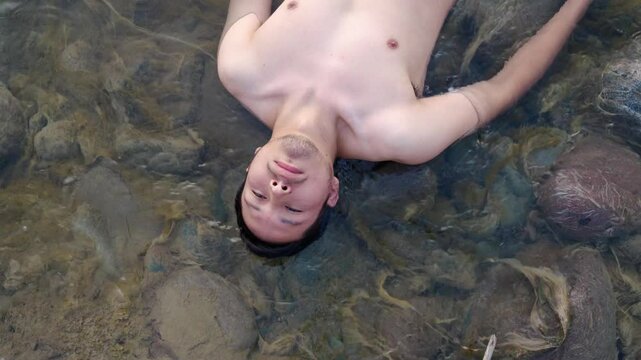 Hombre acostado de espaldas flotando en un r&iacute;o cristalino con los ojos cerrados. Su expresi&oacute;n transmite paz y relajaci&oacute;n. El agua clara revela las piedras del fondo, creando una atm&oacute;sfera natural