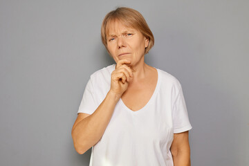 A woman with short hair wearing a white shirt stands against a gray background, looking pensively...
