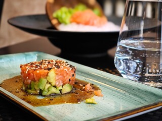 Exquisite salmon tartare on a blue plate with a refreshing glass of water and a background of colorful sushi creations