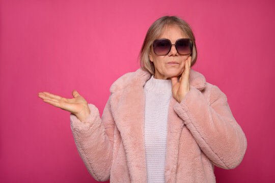 An older woman showcases her fashionable side with oversized sunglasses and a fluffy coat while playfully gesturing against a bright pink backdrop, highlighting her chic style