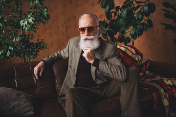Mature gentleman with striking beard posing elegantly on a leather sofa in a studio adorned with...
