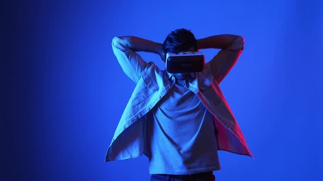 Smart gamer wearing VR goggles while holding sword and fencing in virtual world. Attractive male model playing action game while standing at blue background and accessing in metaverse. Apparatus.