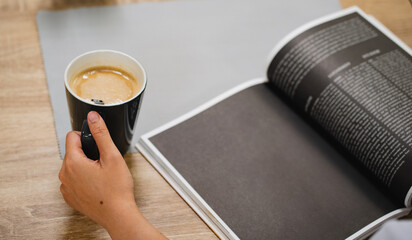 Top of hand holding glass of hot latte  with magazine or book on wooden table in vintage cafe.