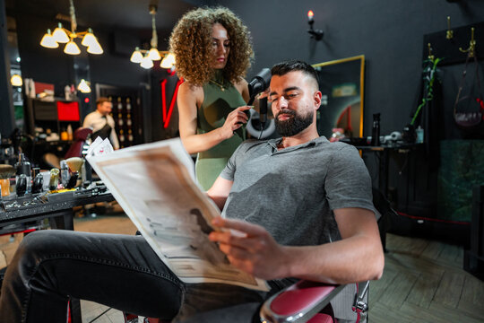 Hairdresser expertly drying man's hair with a hairdryer while he relaxes in a barbershop chair, reading a newspaper and enjoying a well deserved pampering session