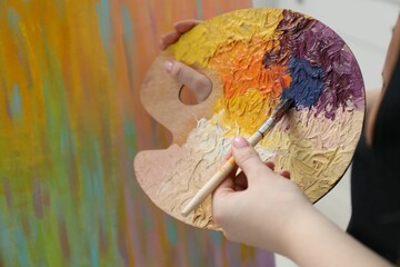 Woman with paintbrush mixing paints on palette near picture, closeup