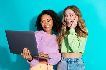Two young cheerful friends using a laptop, enjoying together in colorful casual outfits with a turquoise background
