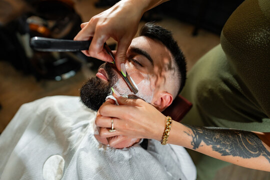 Female barber carefully shaving a male customer's beard with a straight razor, providing a traditional and precise grooming experience in a professional barbershop setting