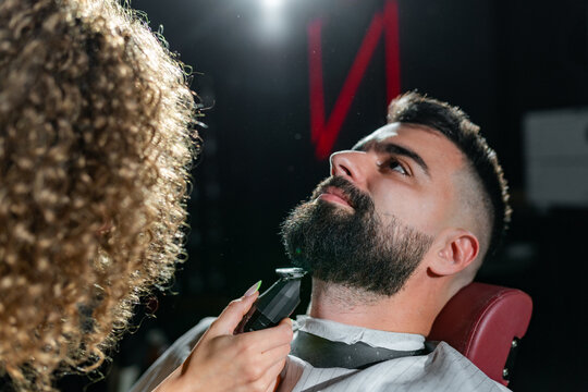 Professional female barber shaving a relaxed male customer's beard with an electric razor in a modern barbershop, providing a stylish and precise grooming experience