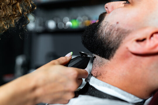 Female barber carefully trimming a male customer's beard with an electric razor, providing a professional and precise grooming experience in a modern barbershop