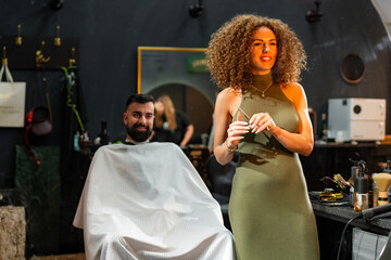 Female hairdresser holding scissors, preparing to cut hair for a male customer seated comfortably...