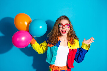 Young woman celebrating with colorful balloons wearing a trendy vibrant outfit and glasses on a blue background.