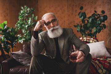 Stylish elderly man with a beard in a retro interior holding a drink and sitting in an elegant and...