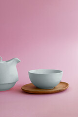 There is a white cup on a wooden saucer on a pink background, and a white teapot in the background. The concept of tableware for tea and coffee.