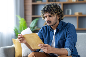 Man reading bad news, received letter in postal envelope. Upset sitting at home on sofa in living...
