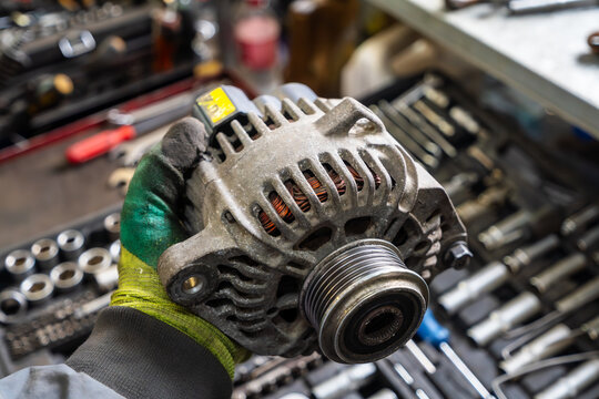 Mechanic wearing gloves holds used car alternator in workshop surrounded by tools and repair equipment.