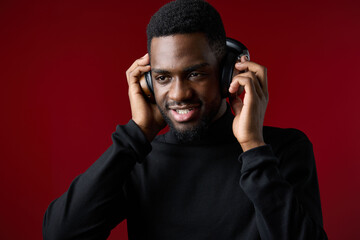 Happy young Black man enjoying music with headphones in a stylish black outfit on a vibrant red...