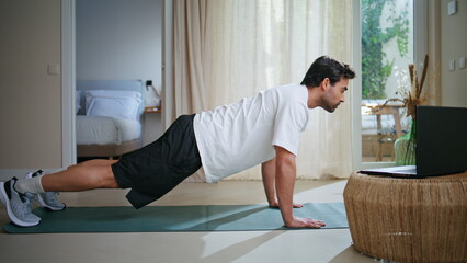 Man performing plank position on yoga mat browsing laptop video. Male athlete