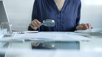 Businesswoman analyzing financial data with magnifying glass and calculator, performing accounting...