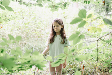 A little cute girl in a T-shirt smiling stands in the middle of a clearing in the forest illuminated by the sun's rays. The concept of children's happiness in nature © PeterPike