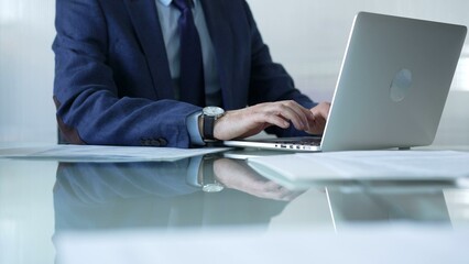 Businessman in blue suit is typing on laptop at glass desk while sitting in bright office, showcasing productivity and modern work environment. Businesspeople concept