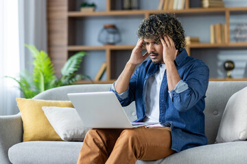 Upset man sitting on sofa in living room at home, in apartment. Employee working remotely using...