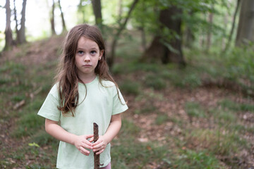 Little cute girl in a t-shirt with a hiking stick on a path in the middle of a summer forest....
