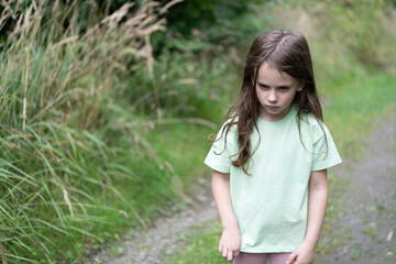 Little sad girl in a t-shirt on a forest road in summer. Concept of a lonely child and sadness