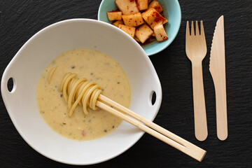 Asian soup with noodles wrapped around disposable chopsticks, served alongside spicy radish kimchi on a black stone surface, accompanied by a wooden fork and knife.
