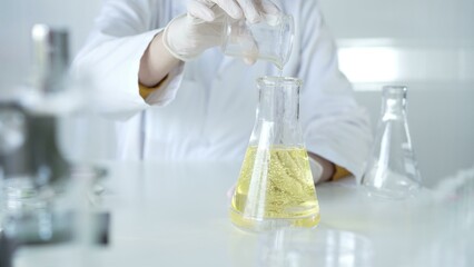 A scientist, wearing a lab coat and white protective gloves, is pouring a yellow oily liquid from one beaker to another in laboratory, close up. Medicine and science concept