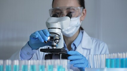 Scientist woman, wearing a lab coat, mask, blue gloves and safety glasses, is adjusting a microscope surrounded by test tubes in laboratory. Medicine and science concept