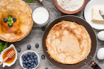 Stack of thin pancakes with blueberries, honey and frying pan with delicious crepes and ingredients for cooking on rustic wooden table. Top view. Delicious breakfast