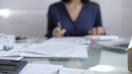 Black calculator, pen and documents are lying on accountant's desk. Businesswoman wearing blue dress is working with documents on the background. Business and audit concept