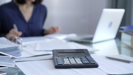 Black calculator, pen and documents are lying on accountant's desk. Businesswoman wearing blue dress is working with documents on the background. Business and audit concept