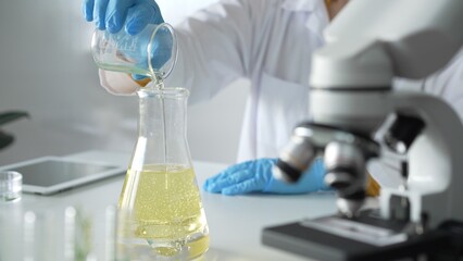 Scientist, wearing blue protective gloves, is holding a beaker with yellow liquid while sitting at the table in laboratory besides microscope, close up. Medicine and science concepts