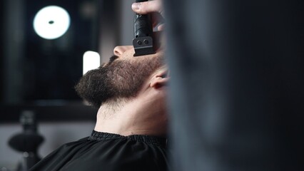 Professional barber trimming client's beard with electric razor, delivering precise styling within...