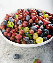 A plate of summer garden berries, gooseberries and blueberries, stands on a gray concrete table.