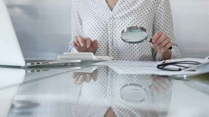 Businesswoman carefully reviewing financial reports with magnifying glass and calculator,...